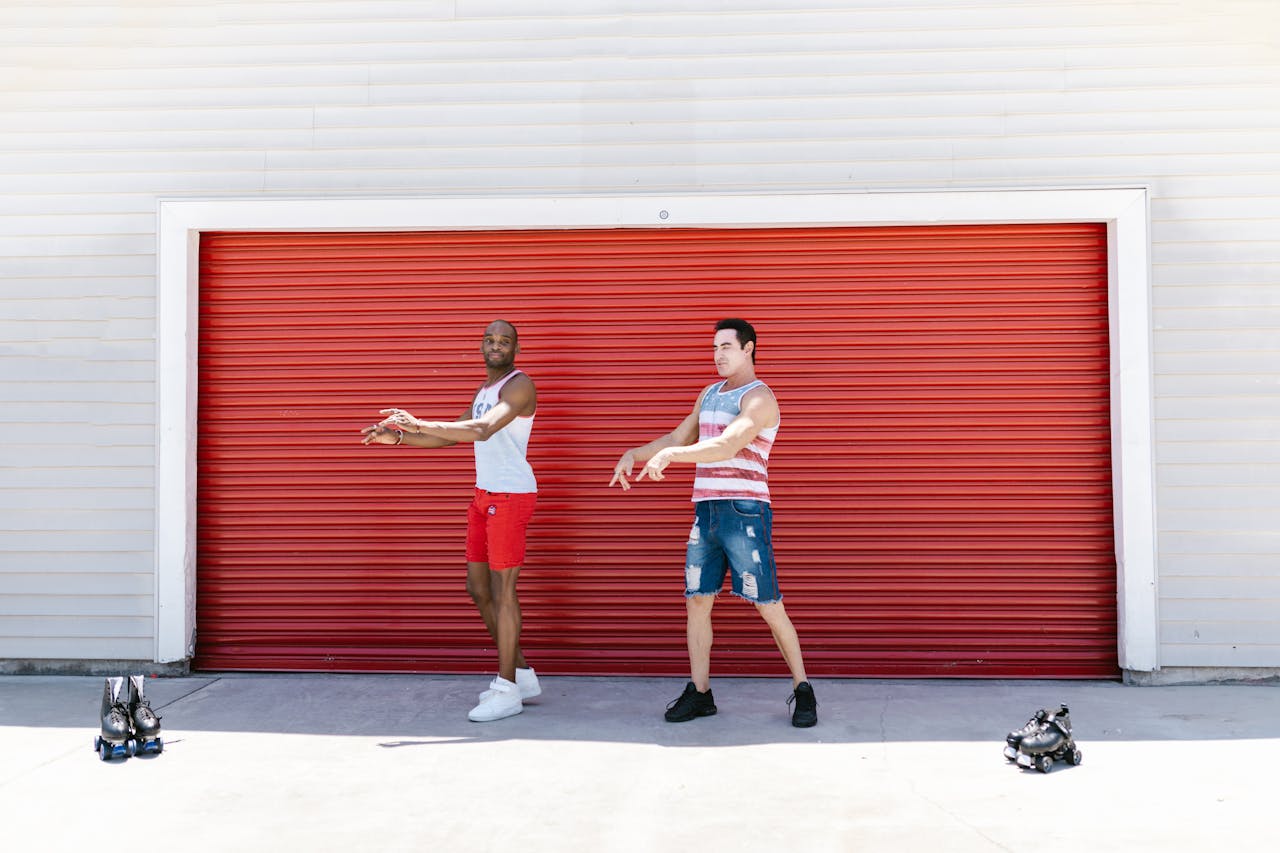 Two men dancing energetically in front of a red garage door, with roller skates beside them.